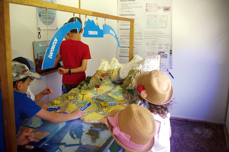 Des enfants rassemblés autour d’une maquette éducative illustrant le cycle de l’eau. Le panneau interactif attire leur attention, offrant une compréhension ludique des processus naturels.