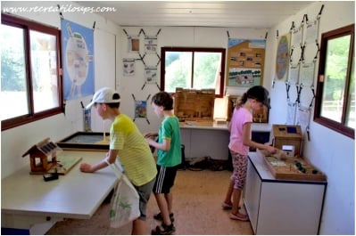 Une scène éducative à l’intérieur d’un chalet où des enfants explorent des maquettes et panneaux interactifs sur l’habitat durable. Une atmosphère studieuse se dégage de leur curiosité.