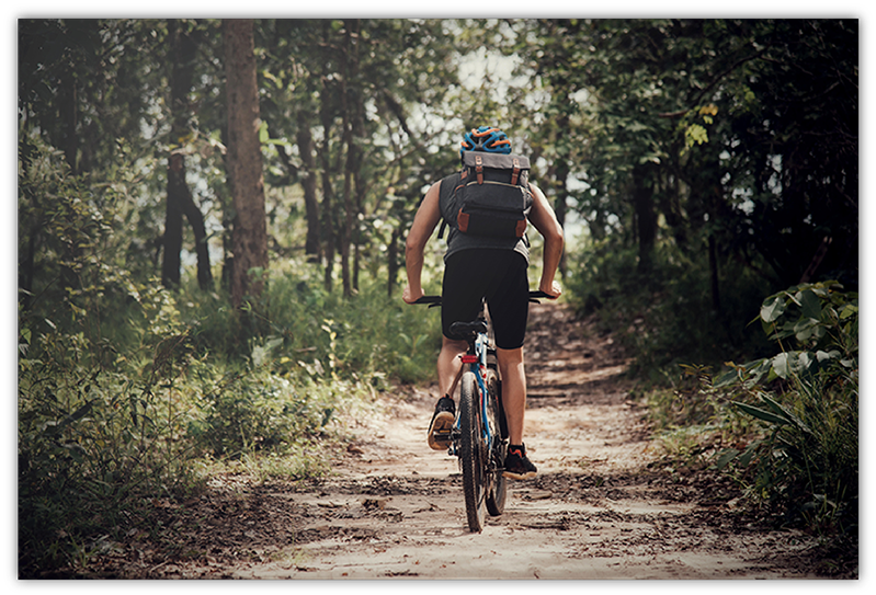 "Un cycliste en tenue sportive parcourant un sentier de randonnée forestière. Il porte un casque et un sac à dos, avançant sur un chemin bordé d'arbres et de verdure, évoquant une expérience immersive en pleine nature."