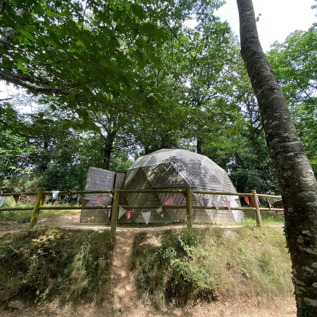 Vue extérieure d'un dôme géodésique en bois entouré de nature, décoré de fanions colorés, servant d'espace d'exposition pédagogique.
