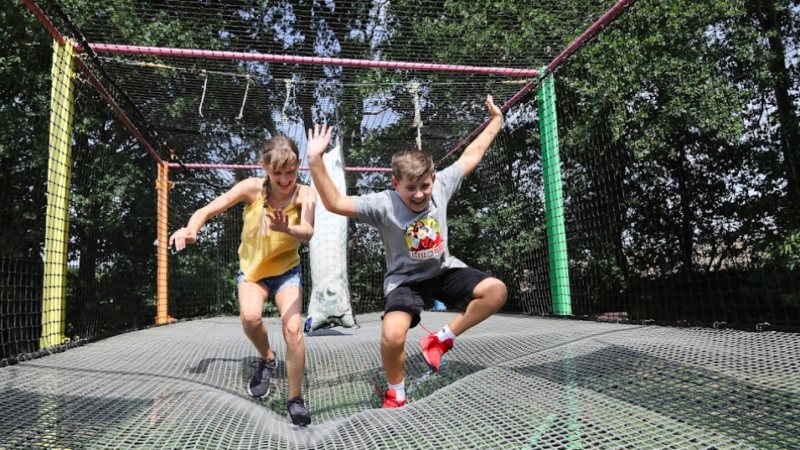 Deux enfants jouent et bondissent dans un filet suspendu, éclatant de rire. La structure est entourée d'arbres verdoyants, et les couleurs vives de la structure créent une ambiance joyeuse et dynamique.