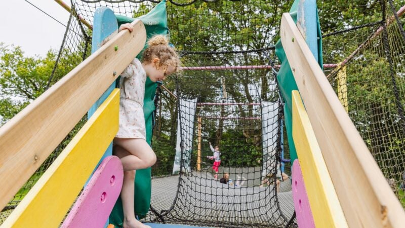 Une petite fille s'apprête à entrer dans le filet suspendu depuis une structure en bois colorée. Derrière elle, d'autres enfants s'amusent à sauter et jouer dans le filet sécurisé, sous le regard bienveillant de leurs accompagnateurs.