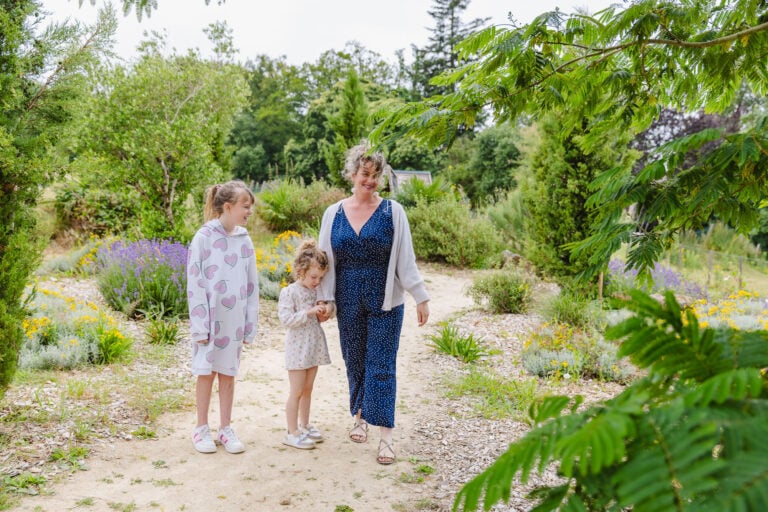 Une promenade familiale à travers un chemin entouré de plantes méditerranéennes. Une femme accompagnée de deux petites filles explore les différentes variétés de plantes, dans un cadre paisible et ensoleillé.