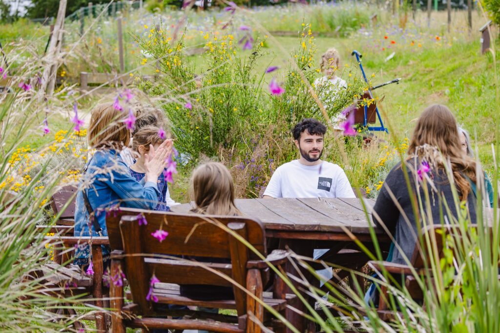 Une famille assise autour d'une table en bois rustique dans le jardin méditerranéen. Entourés de fleurs sauvages et de verdure, les participants profitent d'un moment convivial en plein air.