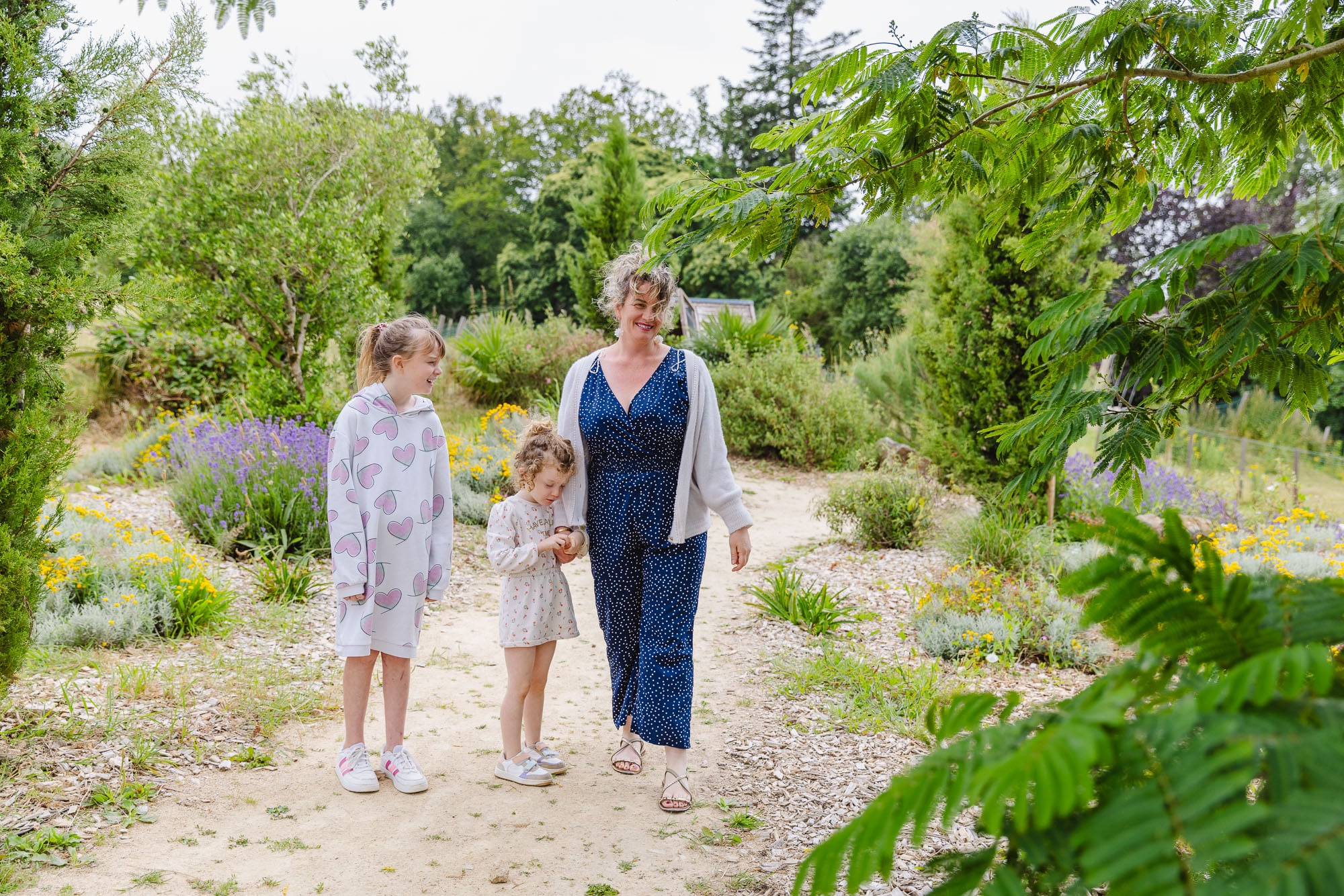 Une promenade familiale à travers un chemin entouré de plantes méditerranéennes. Une femme accompagnée de deux petites filles explore les différentes variétés de plantes, dans un cadre paisible et ensoleillé.