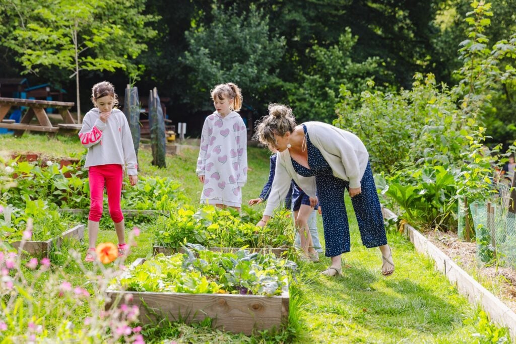 Une maman et un groupe d'enfants inspectent attentivement les carrés potagers. La mère montre les plantes, tandis que les enfants, curieux, examinent les différentes variétés de légumes. L'ambiance est chaleureuse et éducative.