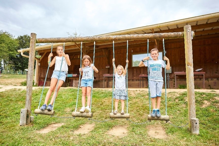 Quatre enfants, souriants et concentrés, se respectent en équilibre sur des rondins suspendus par des cordes. Ils participent à un jeu en plein air devant une structure en bois, mettant à l'épreuve leur agilité et leur coordination.