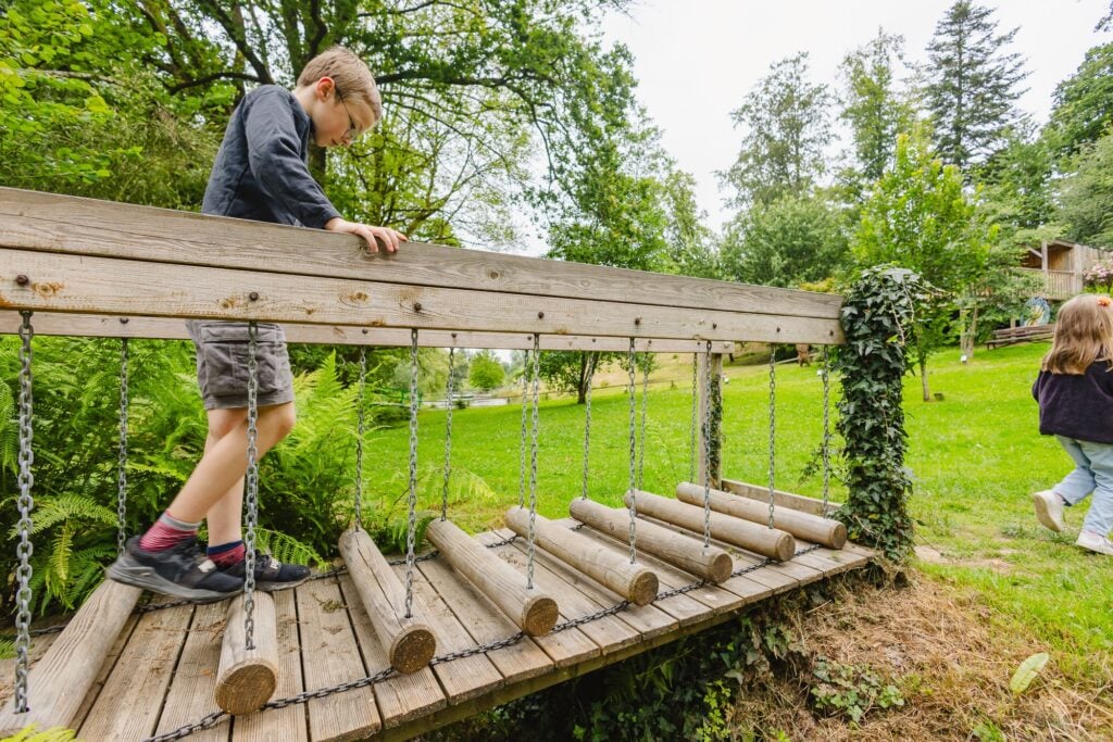 Un jeune traverse un pont suspendu constitué de rondins de bois accrochés par des chaînes, concentré sur son équilibre. La scène est entourée d'arbres et de verdure, créant une ambiance naturelle et ludique.