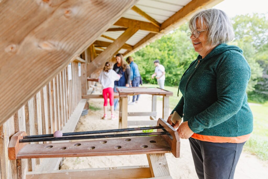 Une femme âgée sourit en jouant avec un jeu en bois où elle pousse une bille à travers un parcours. En arrière-plan, d'autres visiteurs profitent de l'expérience des jeux en plein air.