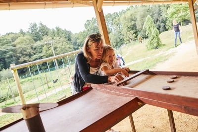 Une mère et sa fille jouent ensemble à un jeu en bois où elles lancent des palets sur une table, dans un cadre convivial et ensoleillé. La guide mère doucement la main de sa fille pour lui apprendre les règles.