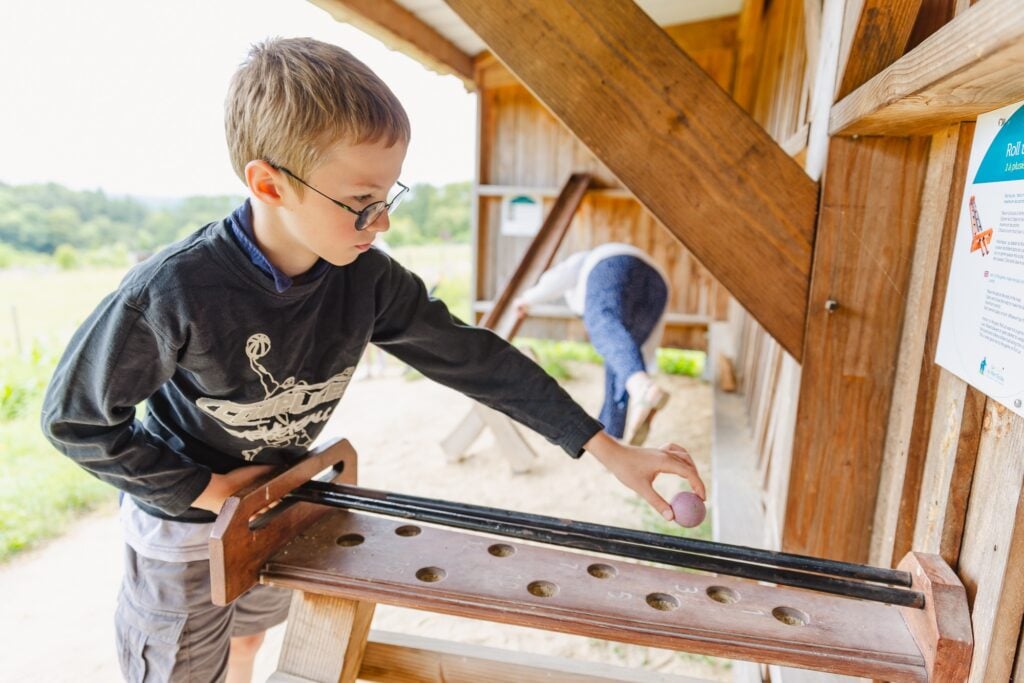 Un jeune garçon concentré déplace une bille en bois sur un parcours incliné, explorant les subtilités du jeu. Le décor rustique et naturel ajoute une ambiance chaleureuse à l'activité.