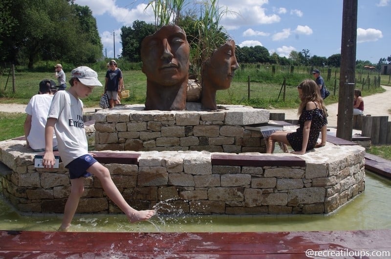 Des enfants s'amusant dans une fontaine entourée de pierres taillées, avec une sculpture artistique représentant deux visages végétalisés au centre, dans un cadre naturel et ludique.
