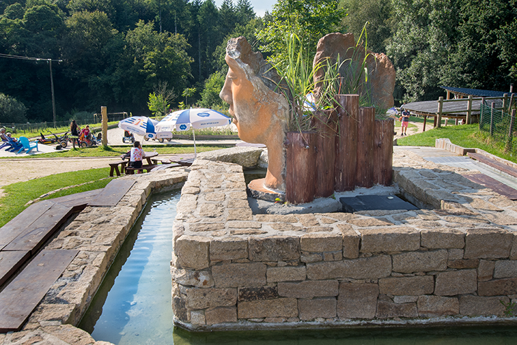 Vue arrière de la fontaine ornée d'une sculpture de deux visages végétalisés, entourée d'un bassin en pierre taillée, avec un espace détente et une ambiance champêtre en arrière-plan.