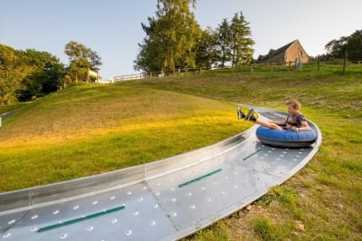 Un garçon glisse avec enthousiasme sur une bouée dans un toboggan métallique incurvé, spécialement conçu pour des descentes sur billes. La pente herbeuse en arrière-plan est baignée par une lumière dorée, avec des arbres et une maison en pierre ajoutant une touche pittoresque à l'environnement naturel.