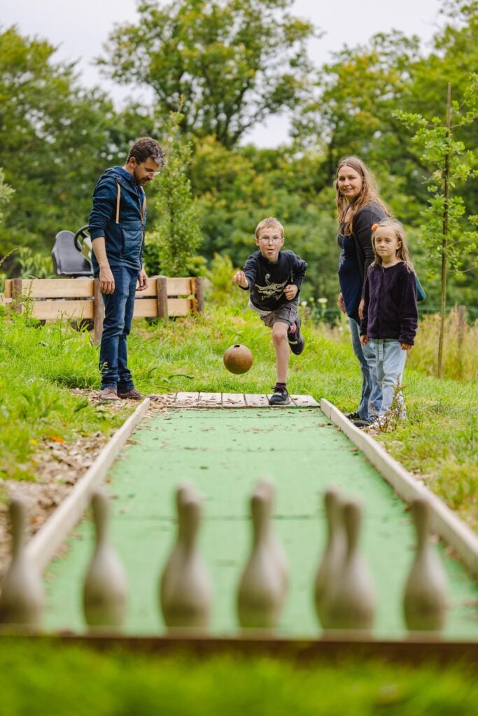Un jeune garçon attentif, regardant la boule de bowling rouler sur la piste verte, tandis que sa famille observe derrière lui, dans un cadre verdoyant et convivial."