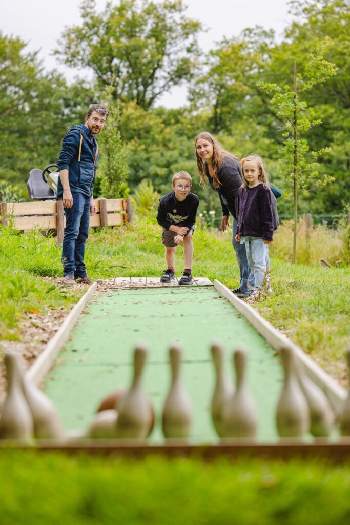 Un garçon en pleine action, lançant une boule de bowling en bois avec énergie, encouragé par sa famille qui profite de ce moment ludique dans un décor bucolique."