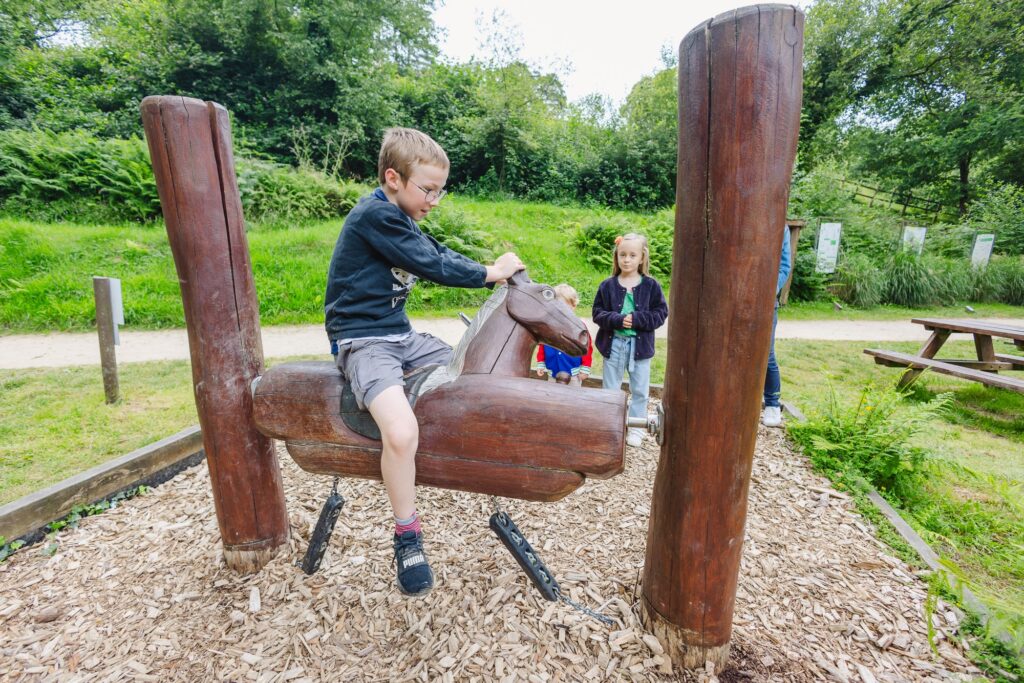 Un jeune garçon joue en équilibre sur un cheval en bois sculpté, entouré de poteaux de soutien. En arrière-plan, deux enfants observateurs avec curiosité. Le cadre naturel est verdoyant, avec un sentier et une table de pique-nique visibles à proximité.
