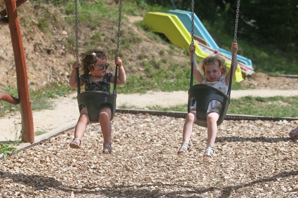 Deux tout-petits se balancent sur des sièges sécurisés dans une aire de jeux en plein air, entourés de bois et d'un toboggan en arrière-plan.