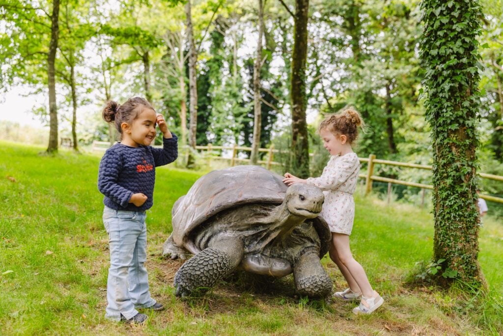 Deux petites filles interagissent joyeusement avec une sculpture réaliste d'une tortue géante. L'une d'elles caresse la carapace tandis que l'autre sourit, créant une scène empreinte de curiosité et de complicité, dans un cadre naturel verdoyant.