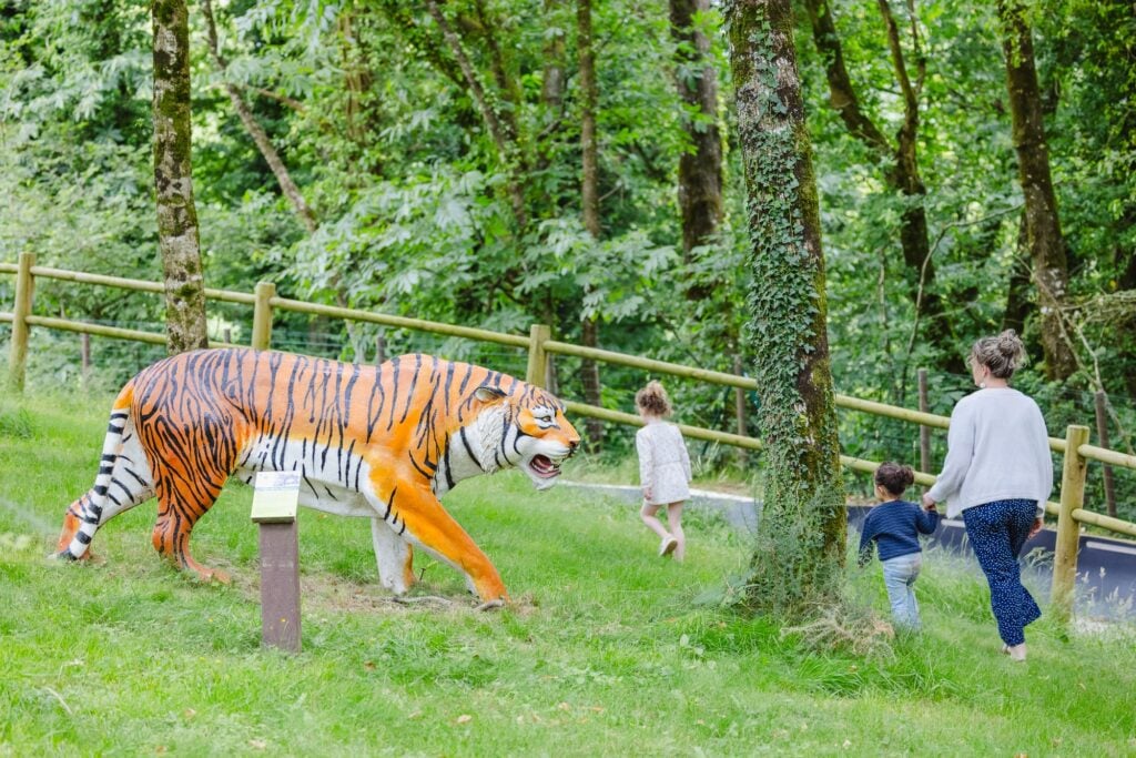 Une famille passe devant une statue colorée d'un tigre réaliste, dans une clairière bordée d'arbres. Les enfants semblent captivés, tandis que la mère accompagne la scène, offrant une expérience immersive dans un décor ludique et éducatif.