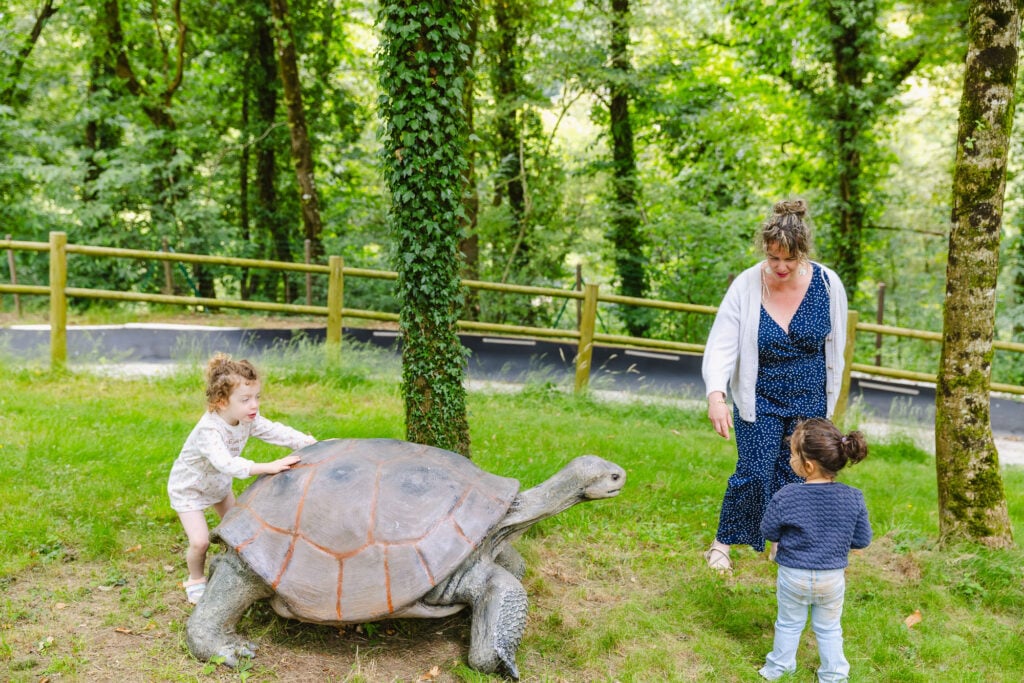 Une mère et deux petites filles s'approchent d'une statue imposante de tortue géante. Tandis que l'une des filles touche la carapace avec émerveillement, l'autre observe avec curiosité, encadrées par un environnement boisé et paisible.