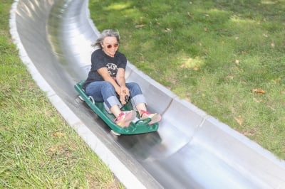 Une femme dévalant une piste de luge d'été en acier, affichant un sourire de plaisir, dans un environnement verdoyant et ensoleillé.