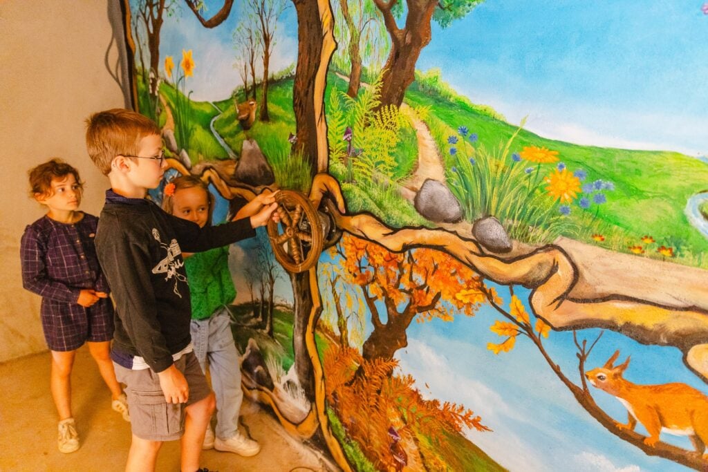 Un groupe d'enfants manipule un mécanisme mural à l'intérieur de la Maison des Hobbits, explorant un décor illustré avec des paysages féériques et des animaux.