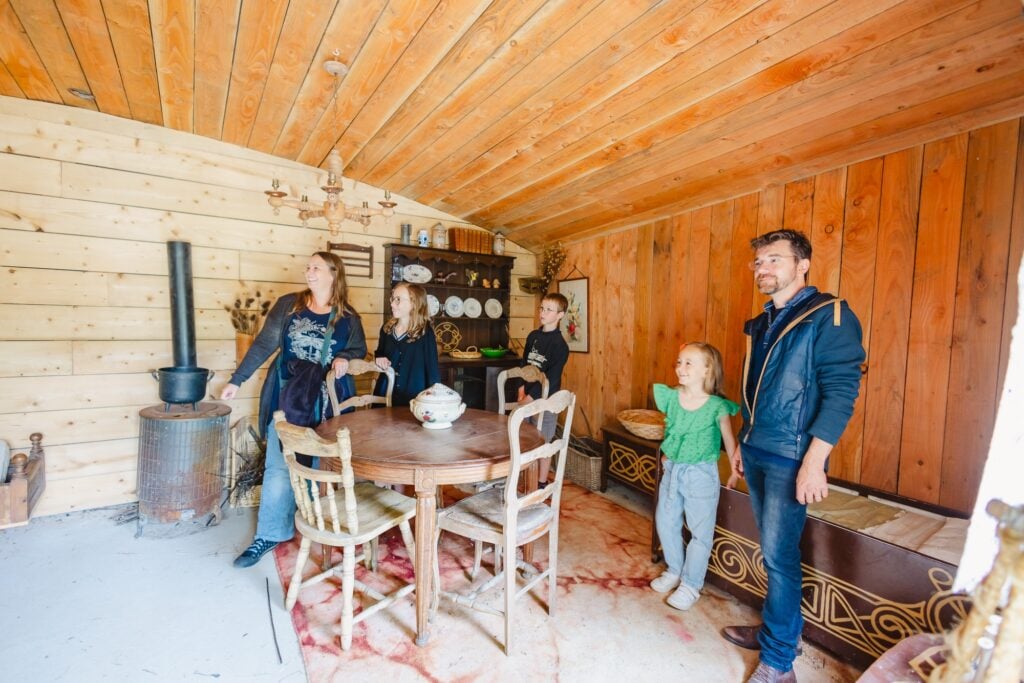 Une famille observe avec intérêt l'intérieur de la Maison des Hobbits, découvrant une table en bois rustique et des éléments décoratifs rappelant une ambiance chaleureuse et traditionnelle.