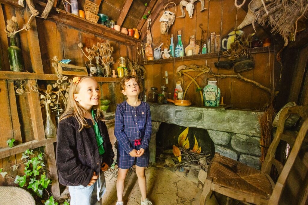 Deux petites filles regardent autour d'elles dans l'intérieur rustique de la maison, rempli de bocaux, d'herbes séchées, et d'objets mystérieux qui évoquent l'univers des sorcières.