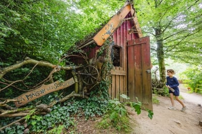 Une jeune fille s'approche de la porte ouverte de la Maison des Sorcières, une petite cabane rouge au style enchanteur entourée de végétation dense. Une pancarte en bois signale "La Maison des Sorcières".