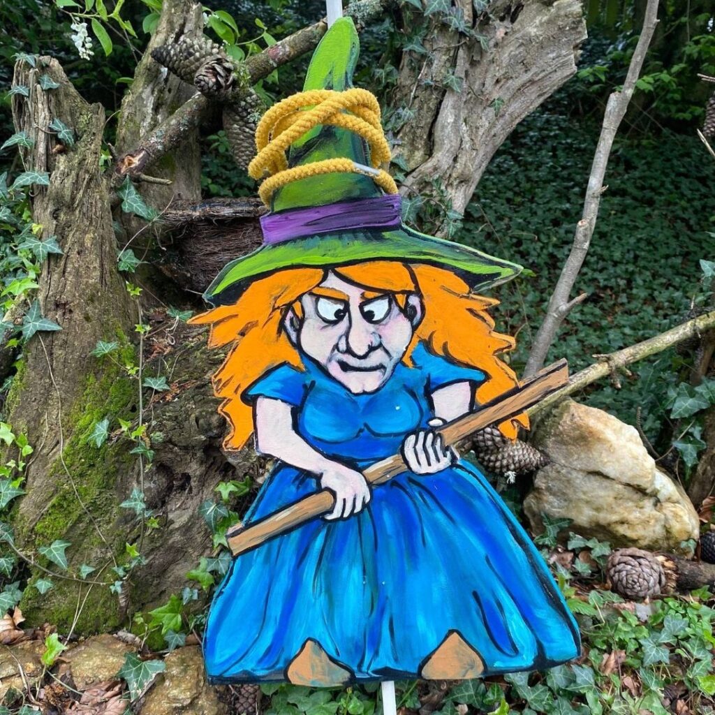 Une deuxième sorcière en bois, peinte avec des cheveux orange et une robe bleue, tient un bâton et accueille un jeu d'adresse pour enfants.