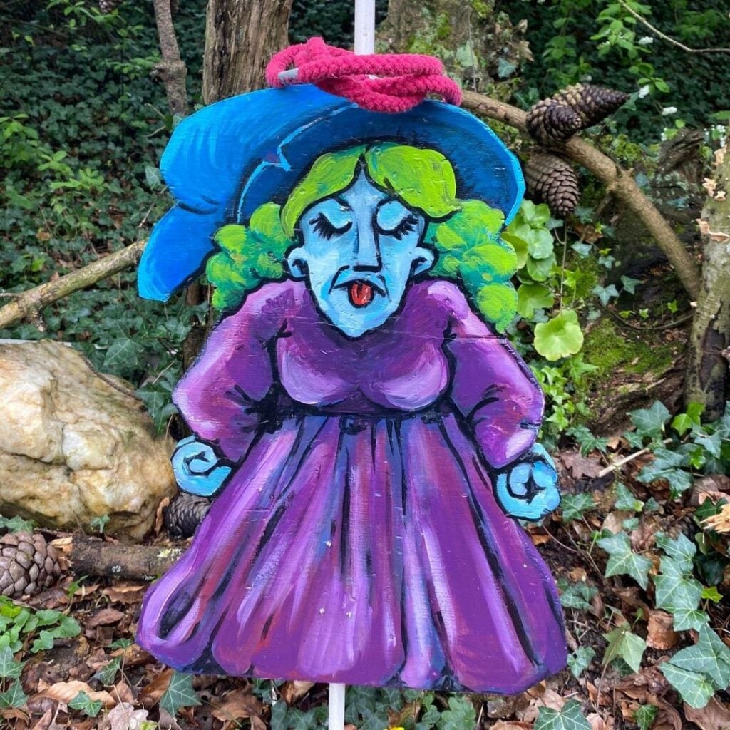 Une sorcière caricaturale peinte en violet avec des cheveux verts et un grand chapeau bleu sert de support ludique pour un jeu d'anneaux, situé au cœur de la forêt.