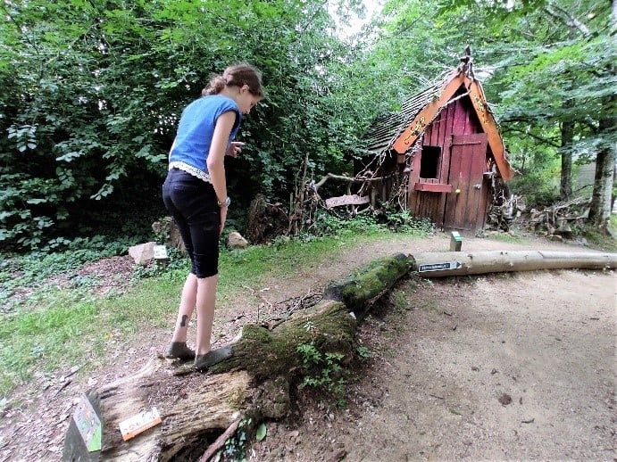 Une jeune fille marche précautionneusement sur un tronc d'arbre disposé comme un jeu d'équilibre, avec la Maison des Sorcières visible en arrière-plan. La scène est entourée d'arbres et de verdure.