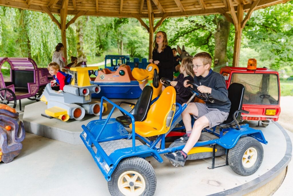 Gros plan sur des enfants s'amusant dans le manège, chacun dans des véhicules colorés tels qu'un chien animé ou une voiture à pédales, sous un pavillon en bois
