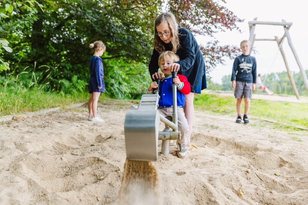 Une petite fille aide un jeune enfant à manier une minipelle dans une zone sablonneuse. Elle guide ses mains sur les poignées pour creuser, tandis qu'en arrière-plan, un garçon observe et une petite fille se tient debout.
