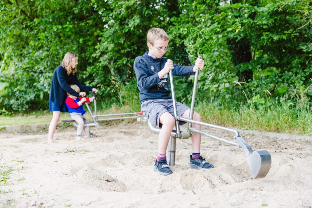 Un garçon utilise une minipelle avec concentration, manipulant les poignées pour creuser dans le sable. À l'arrière, un petit garçon est aidé par une femme sur une autre minipelle. Les enfants sont entourés de verdure et d'arbres, dans un environnement naturel.