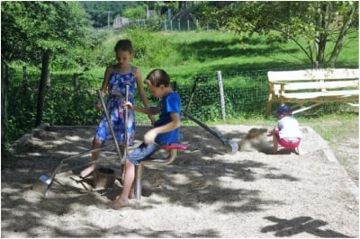 Un garçon et une fille s'amusent avec des minipelles en métal dans un espace de sable. La petite fille utilise les poignées pour manipuler la pelle, tandis qu'un enfant plus jeune joue à l'arrière-plan, assis dans le sable près d'un banc.
