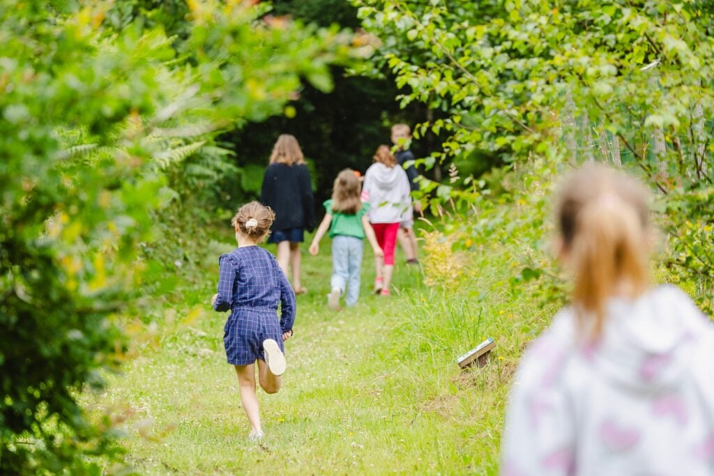 Des enfants courants joyeusement sur un sentier enherbé bordé de verdure, explorant un parcours naturel interactif à la recherche du prochain indice.