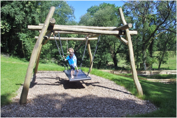 Deux enfants jouent sur un tapis volant suspendu en bois et corde, installé dans un cadre verdoyant. L'un des enfants est debout, tenant fermement les cordes, tandis que l'autre s'assied, profitant du balancement. Le sol est recouvert de copeaux de bois, ajoutant une ambiance naturelle.