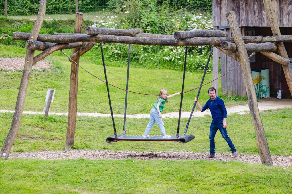 Une petite fille se balance debout sur un tapis volant en bois suspendu, tenant une corde pour garder son équilibre, tandis que son papa, debout à côté, lui donne un léger élan. Le cadre naturel verdoyant et le sol recouvert de copeaux de bois ajoutent une ambiance champêtre et ludique.