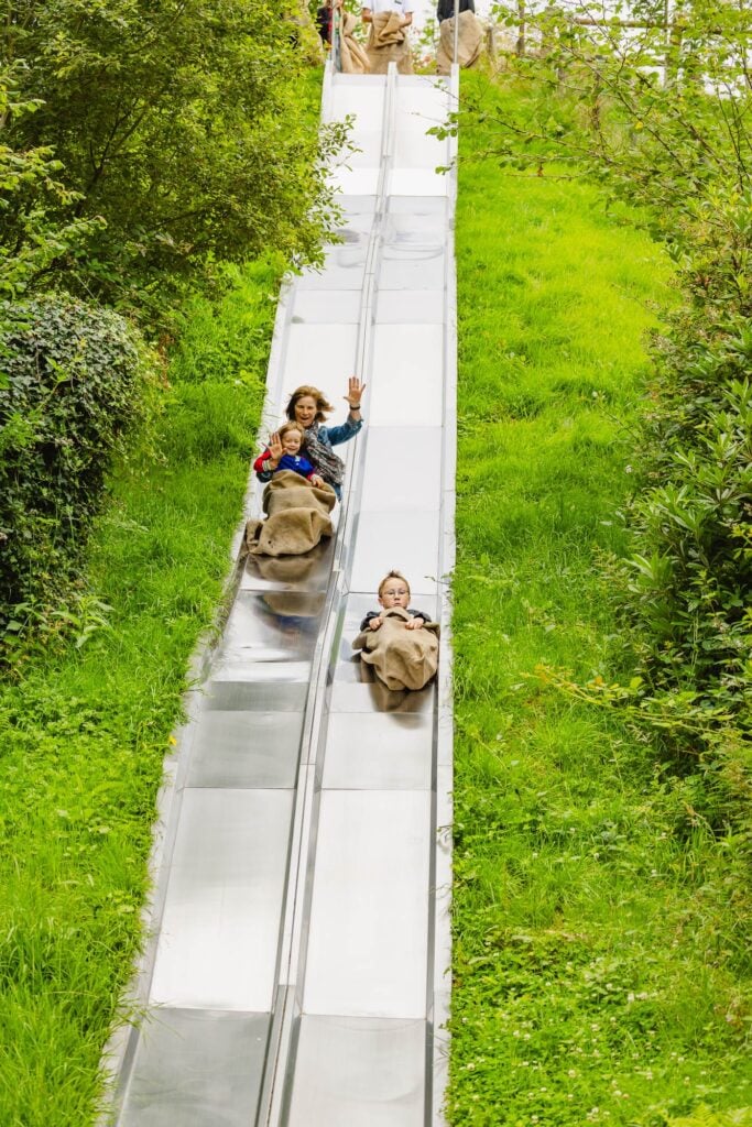 Une grand-mère et son petit-fils dévalant une piste de toboggan géant côte à côte avec un autre garçon, dans un cadre verdoyant et ludique.