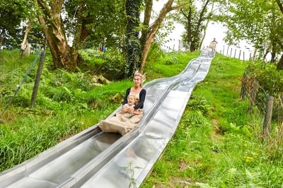 Une femme et un enfant glissant ensemble sur un toboggan géant en acier, entourés d'une nature verdoyante, partageant un moment de joie et de complicité.