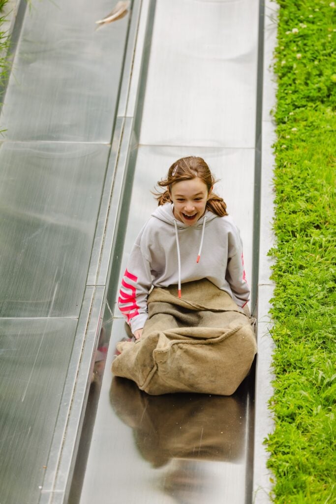 Une jeune fille descend un toboggan géant en acier avec un grand sourire, installée confortablement dans un sac de jute, entourée d'une végétation luxuriante.