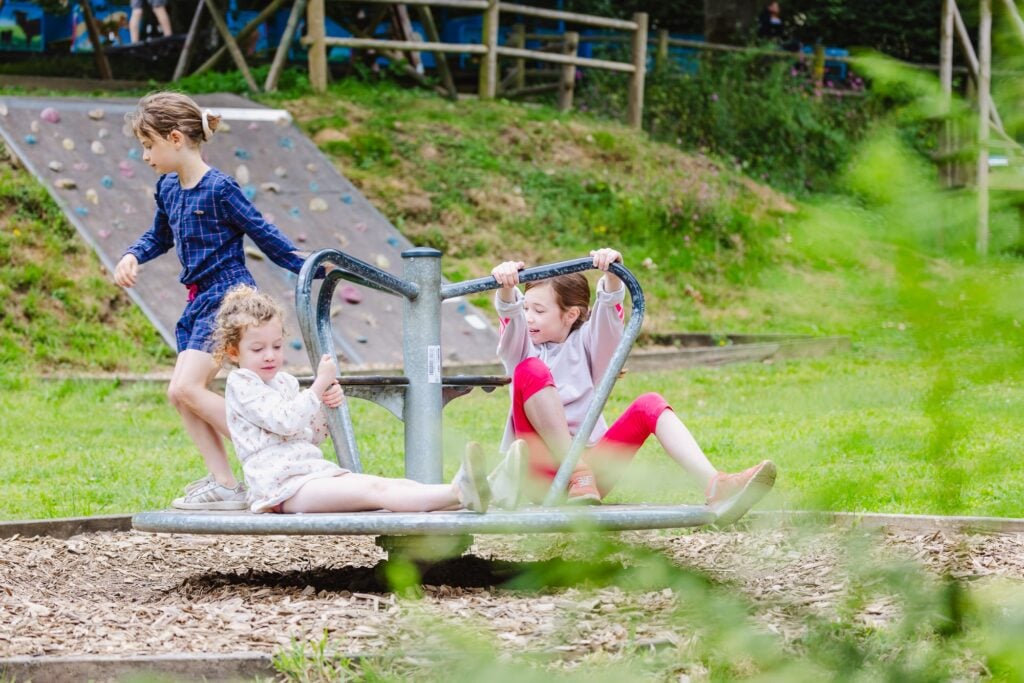 Trois petites filles jouent et rient sur un tourniquet en métal, au milieu d'un espace de loisirs en plein air avec un mur d'escalade visible à l'arrière-plan.