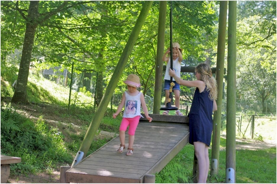 Une petite fille montée sur le siège de la tyrolienne avec l'aide d'une adulte, prête à profiter d'une expérience ludique dans un cadre naturel et ombragé.