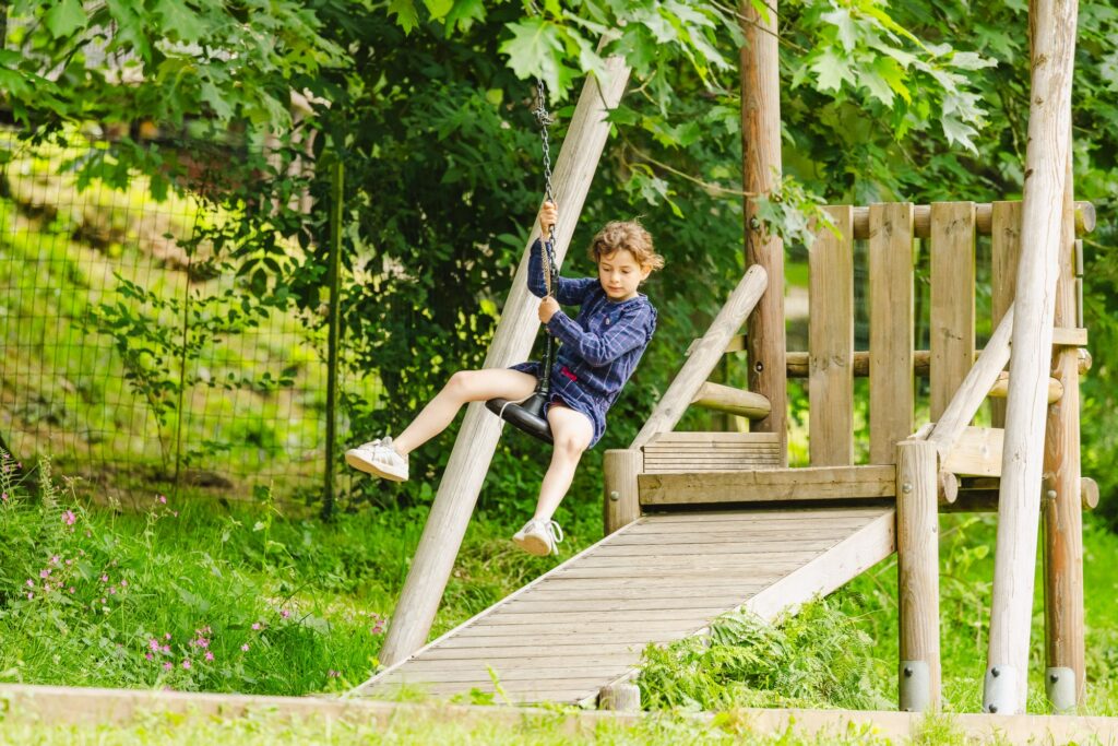Une enfant en pleine descente sur une tyrolienne à double sens, s'amusant sur une structure en bois entourée de verdure, dans un espace de jeu en plein air.