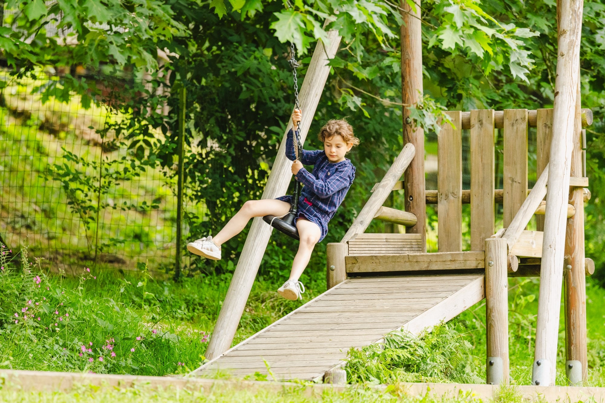 Une enfant en pleine descente sur une tyrolienne à double sens, s'amusant sur une structure en bois entourée de verdure, dans un espace de jeu en plein air.