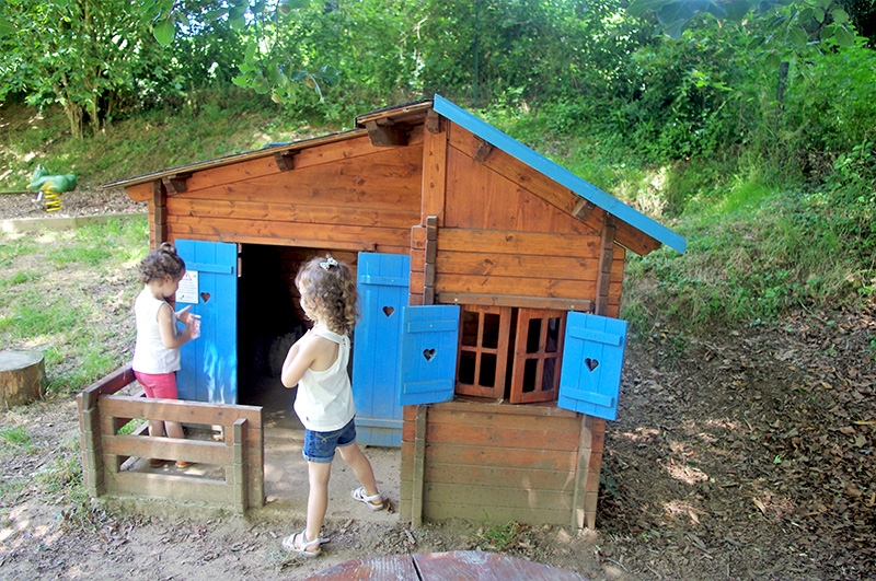 Deux jeunes filles jouent autour d'un charmant chalet en bois. Les volets bleus ornés de cœurs ajoutent une touche accueillante. Ce lieu offre un espace idéal pour les jeux d'imagination en plein air.