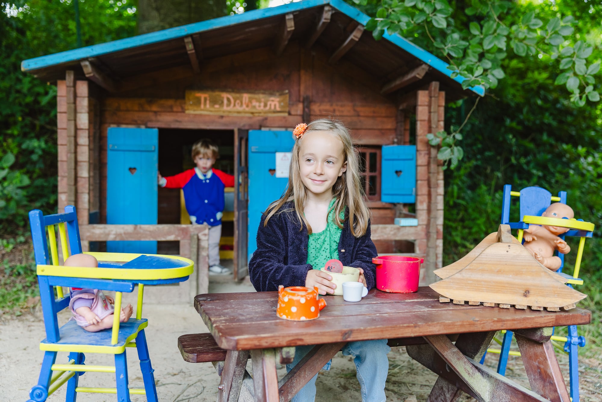 Une petite fille joue à faire semblant de préparer un repas à une table de pique-nique devant un chalet de cuisine miniature. Les accessoires, comme une petite théière colorée, ajoutent une dimension réaliste à ce jeu d'imitation.