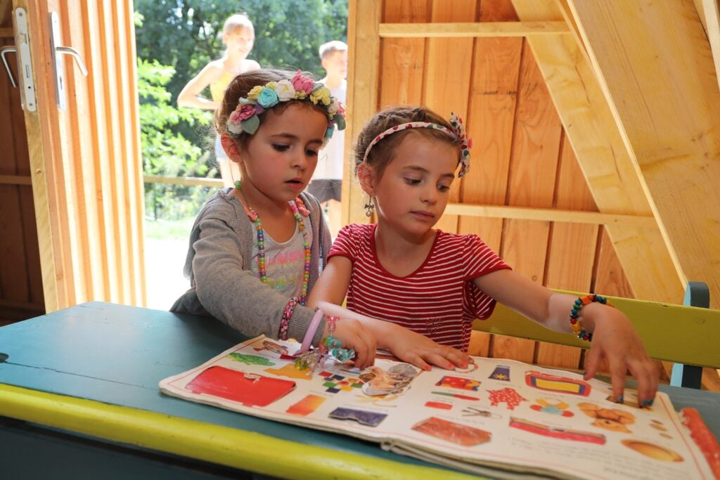 Deux petites filles concentrées dans une activité créative à l'intérieur d'un chalet éducatif. Des livres ouverts devant elles laissent imaginer des jeux autour de l'apprentissage et de la découverte.